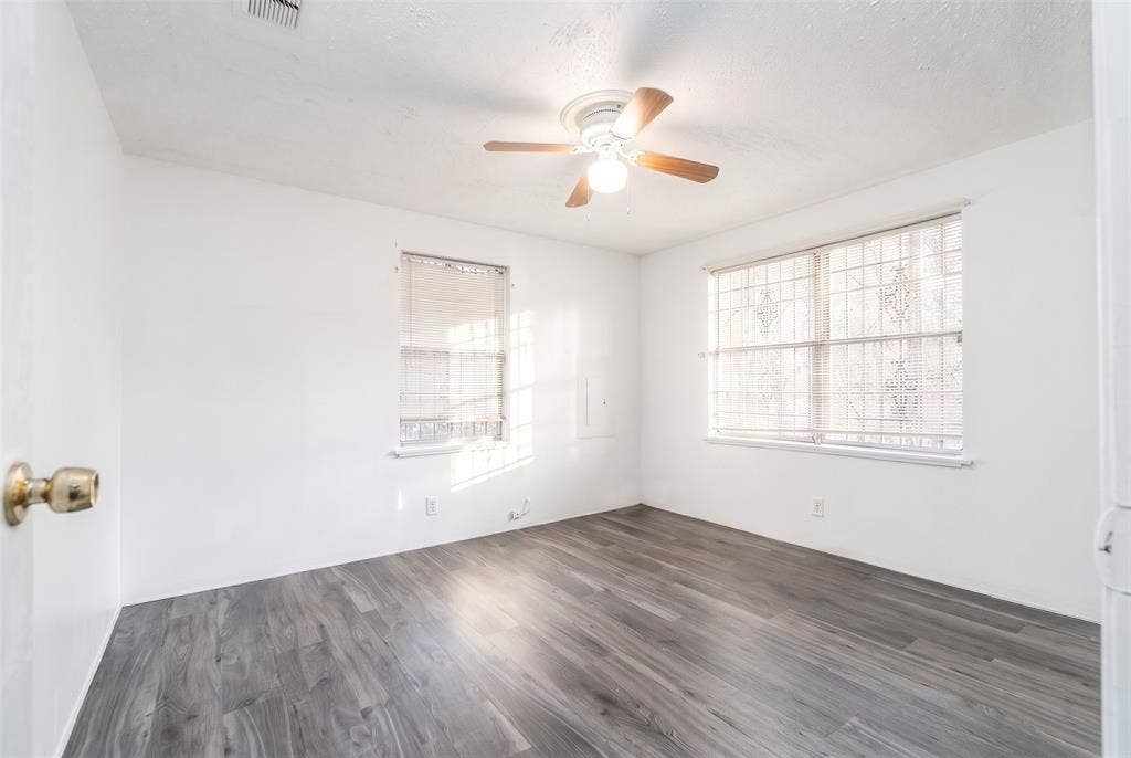 Neutral-toned room featuring wood-finish flooring, a ceiling fan with integrated light fixture, two windows, and security bars