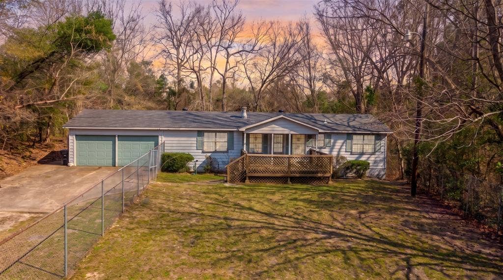 Single-story exterior featuring light-toned siding, attached two-car garage, front wood deck with lattice skirting, concrete driveway, and perimeter chain-link fencing