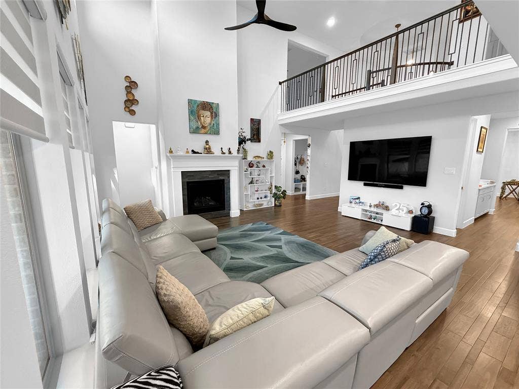 2251 Macy Way, Prosper, TX 75078 High-ceiling interior featuring a dark-tiled fireplace surround, wood-finish flooring, a matte black ceiling fan, and a mezzanine level with a metal railing