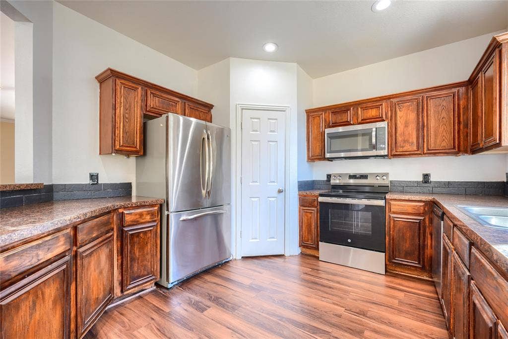 2709 Maci Court, Seagoville, TX 75159 Kitchen featuring wood-finish cabinetry, stainless steel appliances, speckled stone-finish countertops, dark tile backsplash, and wood-finish flooring