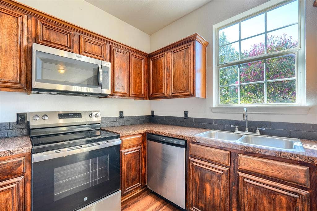 2709 Maci Court, Seagoville, TX 75159 Kitchen featuring stained wood cabinetry, stainless steel appliances, dark tile backsplash, speckled countertops, and a double-basin stainless steel sink under a multi-pane window