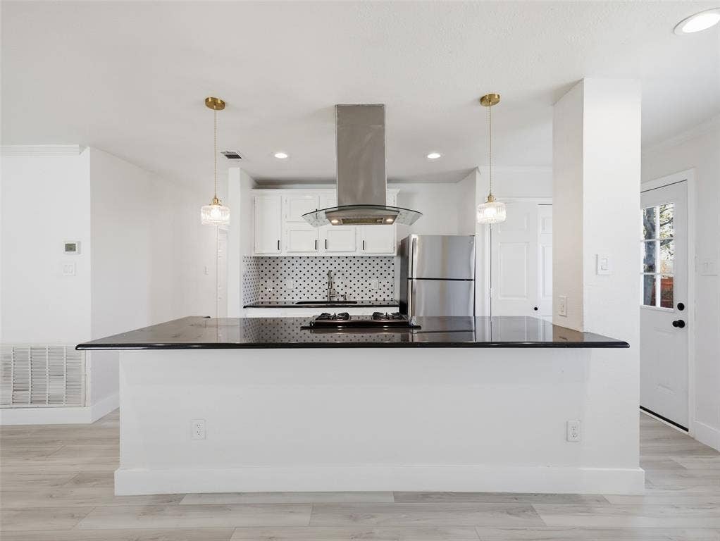 Kitchen featuring a central island with a gas cooktop, dark stone-finish countertop, stainless steel vent hood, white cabinetry, and glass pendant lighting