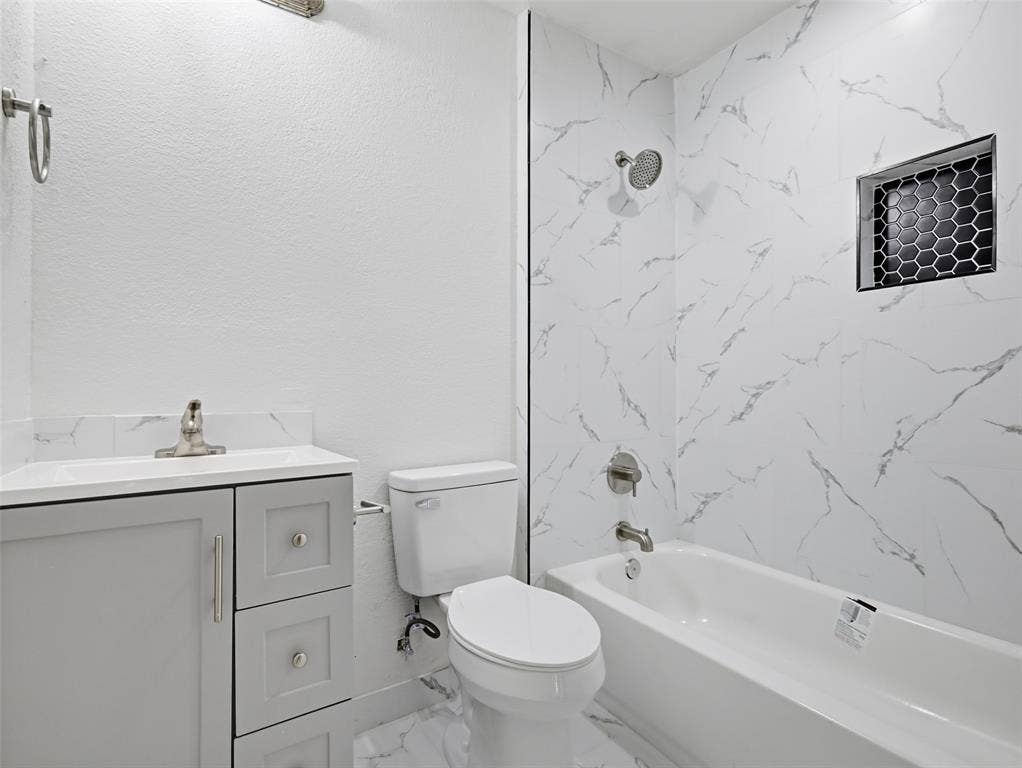 Bathroom featuring a white vanity cabinet with a stone-top integrated sink, bathtub with stone-patterned wall tile, and a decorative hexagonal tile shower niche