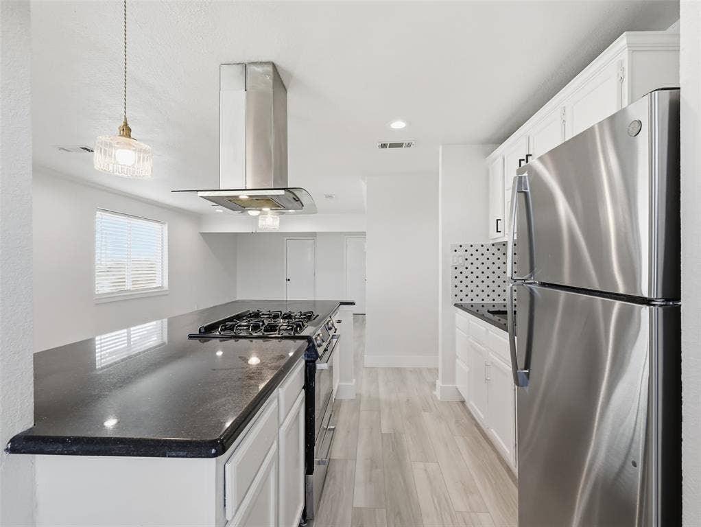 Kitchen featuring white cabinetry, dark stone-finish countertops, stainless steel appliances including a gas range, a modern stainless steel chimney-style range hood, and wood-finish flooring