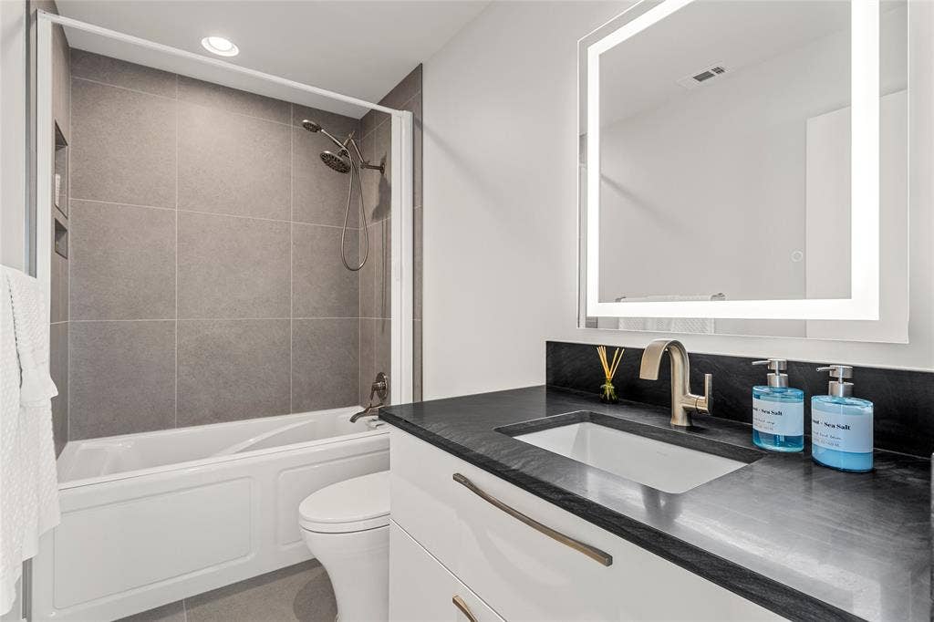 356 Bold Springs Court, West, TX 76691 Bathroom featuring a floating white vanity with dark stone-finish countertop, undermount sink, illuminated wall-mounted LED mirror, and a shower-tub combination with retractable privacy door. 24 inch custom tile work.