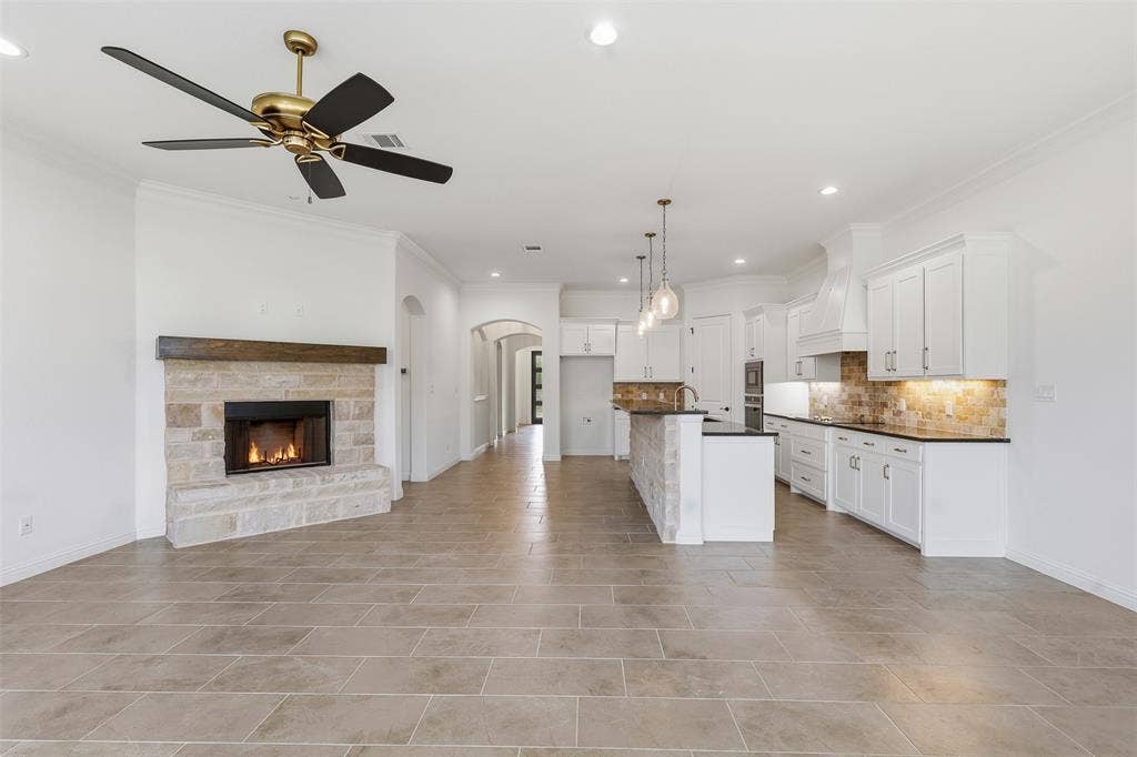 Open-concept interior featuring large-format tile flooring, a floor-to-ceiling stone fireplace with a wood mantel, white cabinetry, dark countertops, and a multi-light pendant fixture over the kitchen island