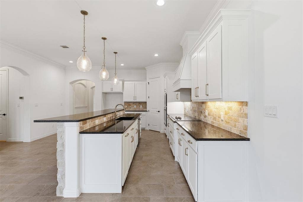 Kitchen featuring white shaker-style cabinetry, dark stone countertops, natural stone tile backsplash, stone-faced breakfast bar, and three glass pendant lights