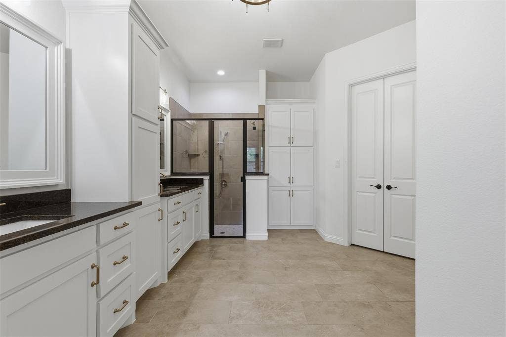 White cabinetry with dark solid-surface countertops, walk-in shower with dark-toned frame, floor-to-ceiling built-in storage, and large-format tile flooring
