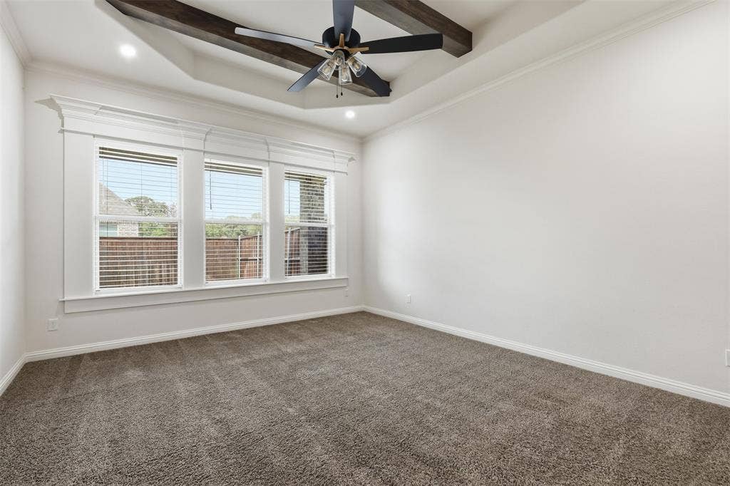 Tray ceiling with exposed wooden beams, center-mounted ceiling fan, neutral wall-to-wall carpeting, and a triple-pane window assembly with detailed millwork trim