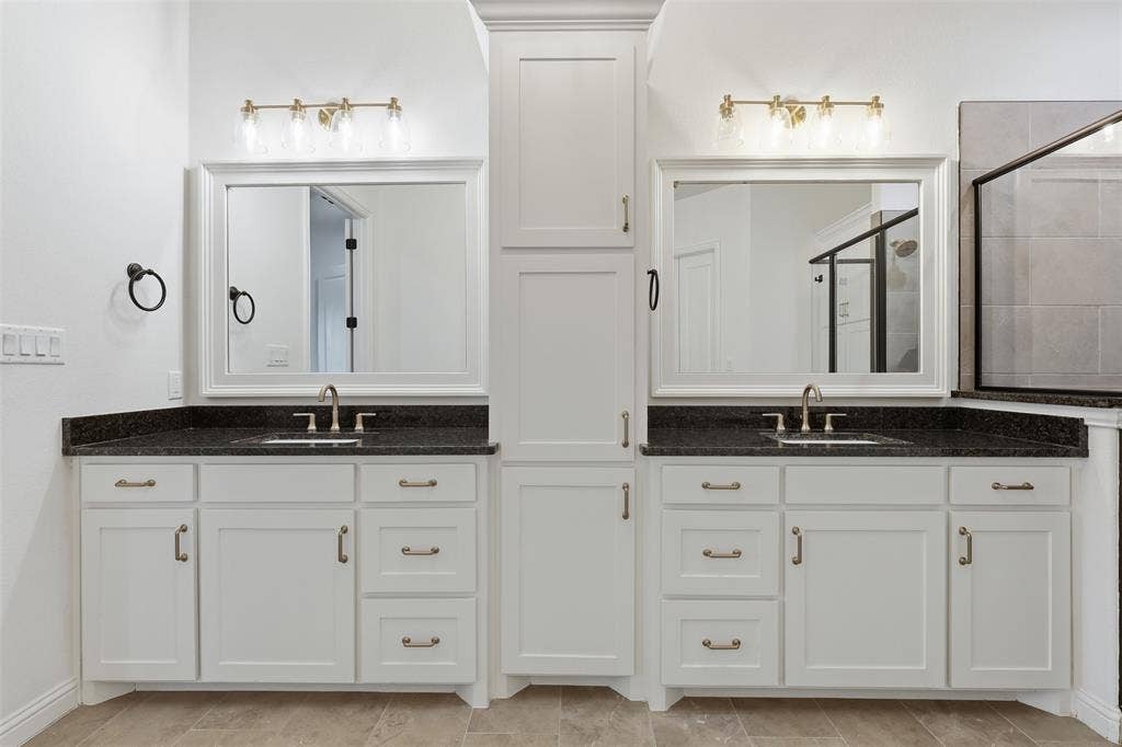 Dual-sink vanity featuring white cabinetry, dark stone-finish countertops, central built-in storage, framed mirrors, and metallic wall-mounted light fixtures