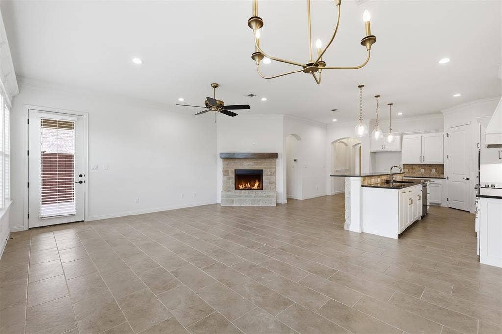 Open concept living area featuring large-format tile flooring, a stone-surround fireplace with a wooden mantel, a kitchen island with an integrated sink, white shaker-style cabinetry, and multi-light chandelier fixtures