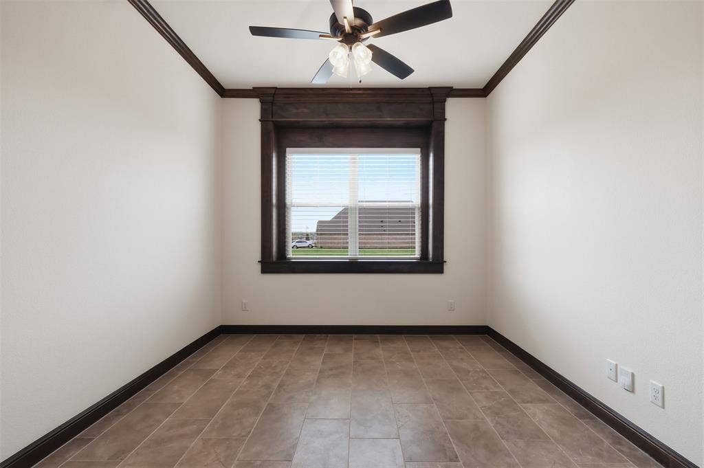 Room featuring large-format tile flooring, dark wood-finish crown molding, ceiling fan with integrated lighting, and a central window framed by matching dark wood-finish casing