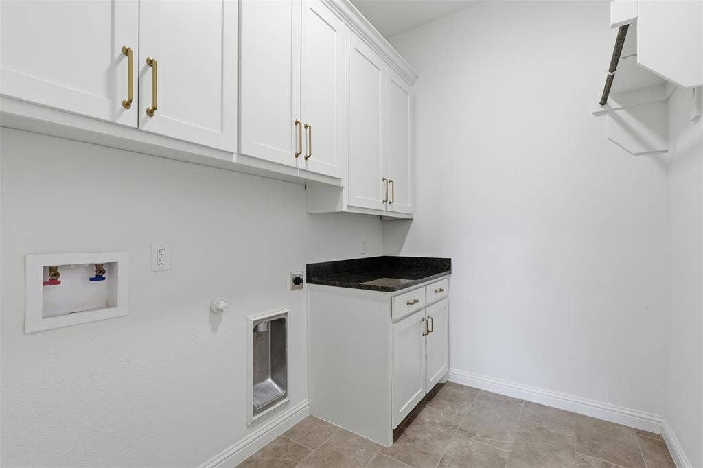 Laundry room featuring white shaker-style cabinetry with brass-tone hardware, a dark stone-surface countertop, an integrated sink, and tile flooring