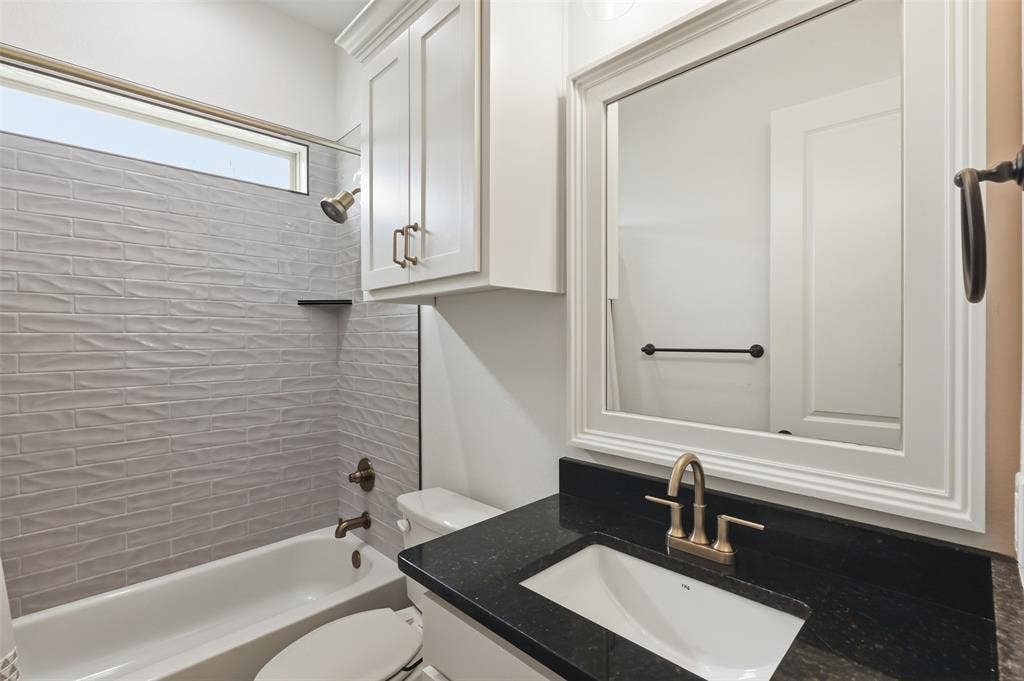 Bathroom featuring a dark stone countertop, integrated sink with a metallic faucet, white cabinetry, a framed mirror, and a tub surround with textured tile