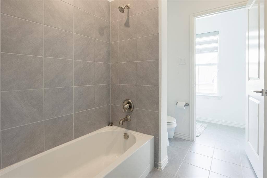 Bathtub featuring full-height gray wall tile, polished metal faucet and shower head, and tile flooring