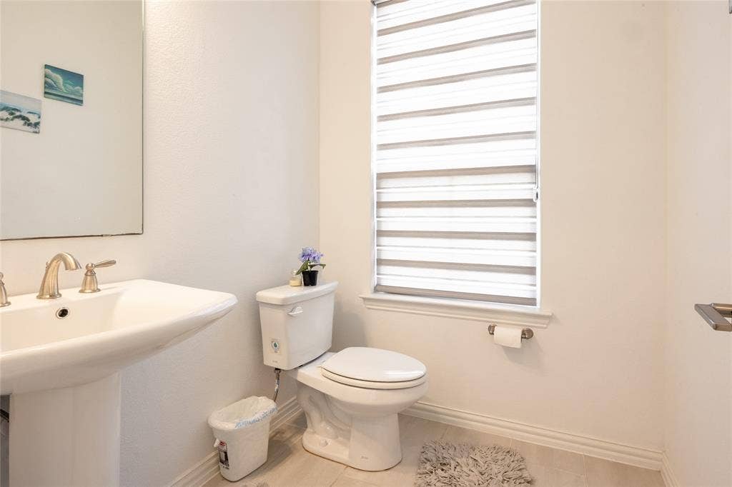 Pedestal sink with polished metal hardware, large wall-mounted mirror, white elongated toilet, and window featuring horizontal light-filtering shades