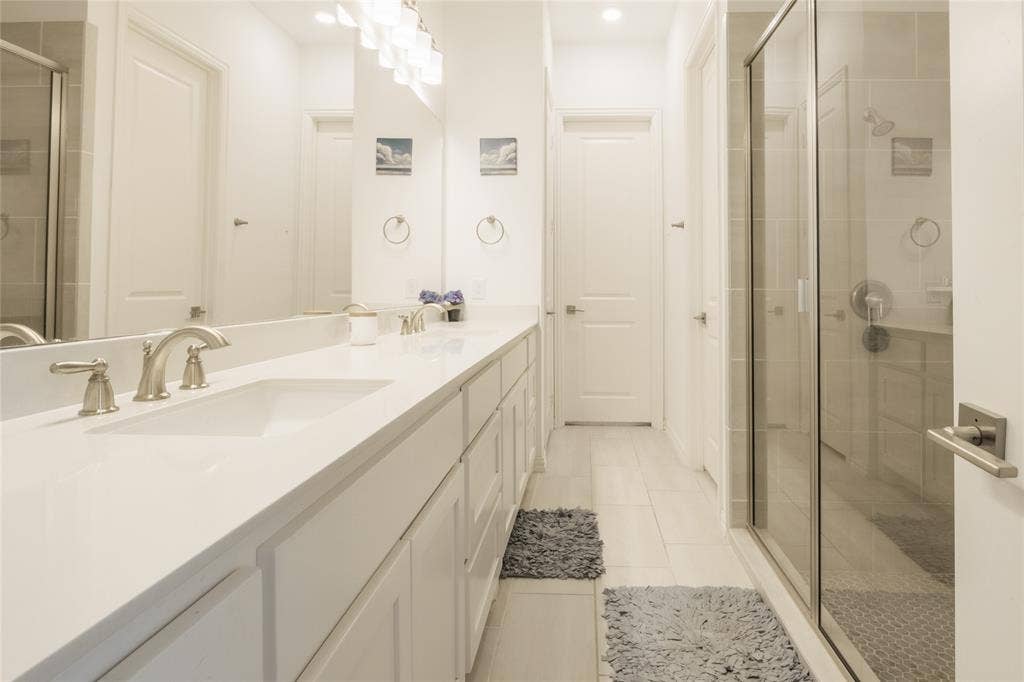 Bathroom featuring a long double vanity with solid surface countertops, dual undermount sinks, white shaker-style cabinetry, and a glass-enclosed shower with tile surround