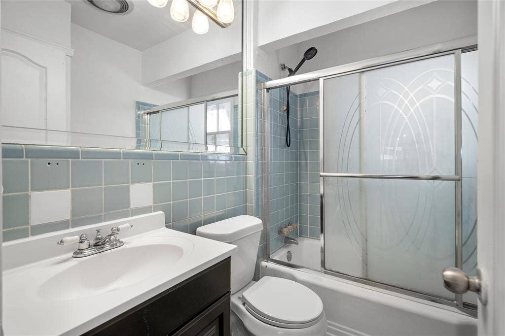 2005 Tisinger Avenue, Dallas, TX 75228 Bathroom featuring a dark-finish vanity with a white integrated sink, full-width wall mirror, shower-tub combination with blue wall tiling, and glass sliding enclosure with etched detailing