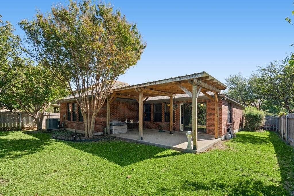 Rear exterior featuring a brick facade, covered patio with a wooden pergola, expansive lawn, and mature trees enclosed by wood fencing