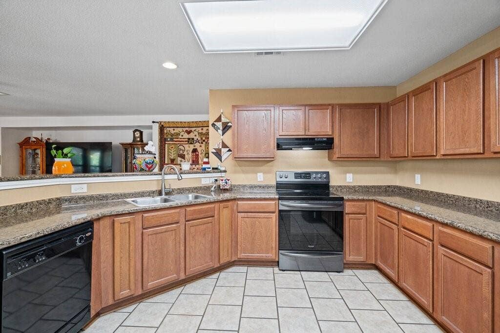 Kitchen featuring wooden cabinetry, stone-finish countertops, black appliances, a double-basin stainless steel sink, and tile flooring