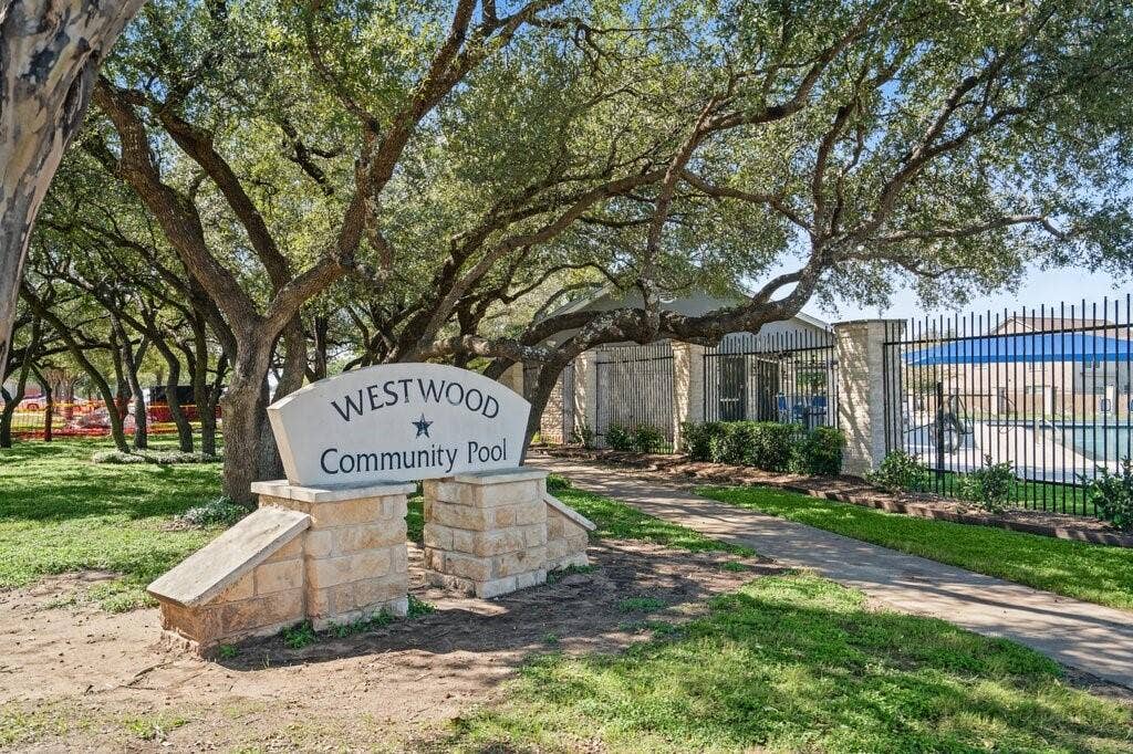 Community entrance featuring a stone monument sign, mature oak trees, paved walkway, and fenced swimming pool area