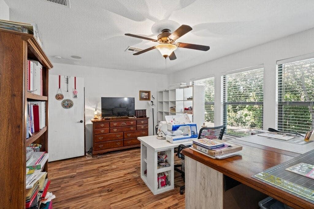 Room featuring wood-finish flooring, three windows with horizontal blinds, and a multi-blade ceiling fan with an integrated light fixture