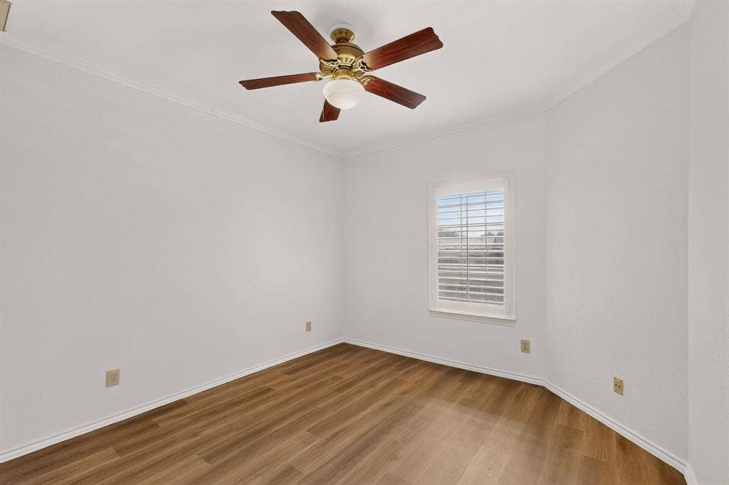 The crisp secondary bedroom features fresh paint, newly installed wood-finish laminate flooring, crown molding, a central ceiling fan with integrated lighting, and a window fitted with interior plantation shutters.