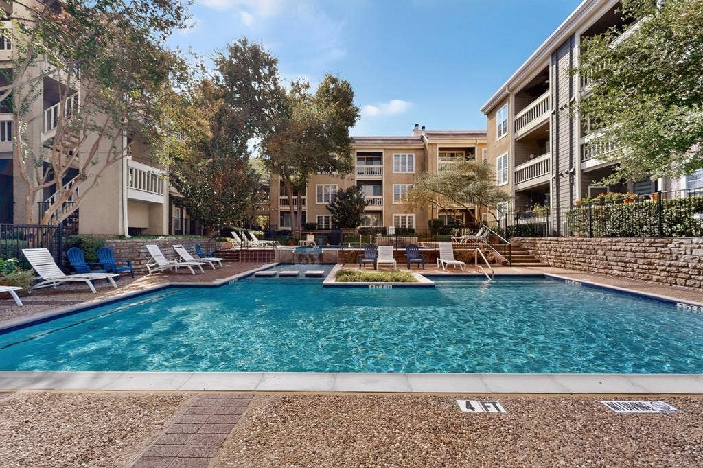 Multilevel stucco exterior condominium complex featuring private balconies with railings, expansive swimming pool with integrated spa, surrounding stone paver decking, community grill and more.