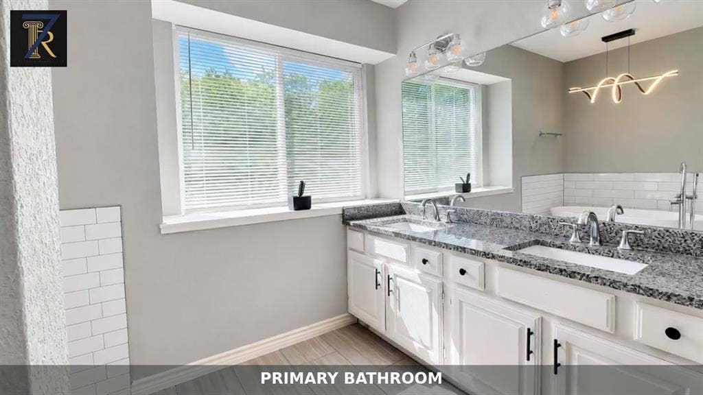 Dual-sink vanity with stone countertop, white cabinetry with matte black hardware, subway tile accents, wood-finish flooring, and sculptural modern lighting