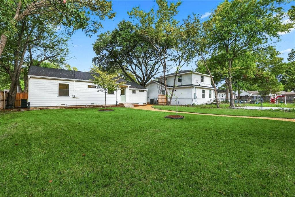 11624 Rupley Lane, Dallas, TX 75218 Expansive backyard featuring a large grass lawn, mature shade trees, a light-toned gravel pathway, and white horizontal siding exteriors