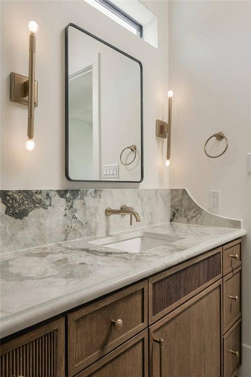 Custom wood vanity with reeded detailing, stone countertop with matching backsplash, undermount sink, brass wall-mounted faucet, and rectangular mirror with dual brass light fixtures