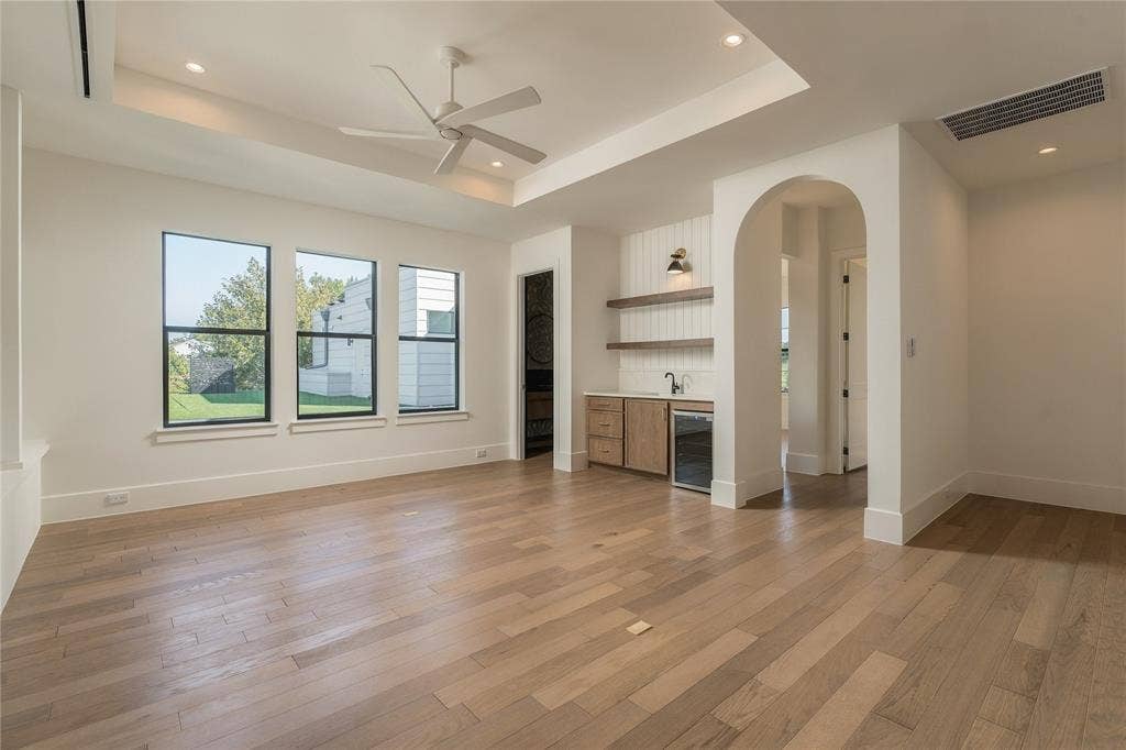 Tray ceiling with integrated recessed lighting, wood-finish flooring, built-in wet bar featuring stone countertops, shiplap backsplash, and open shelving, plus an arched entryway