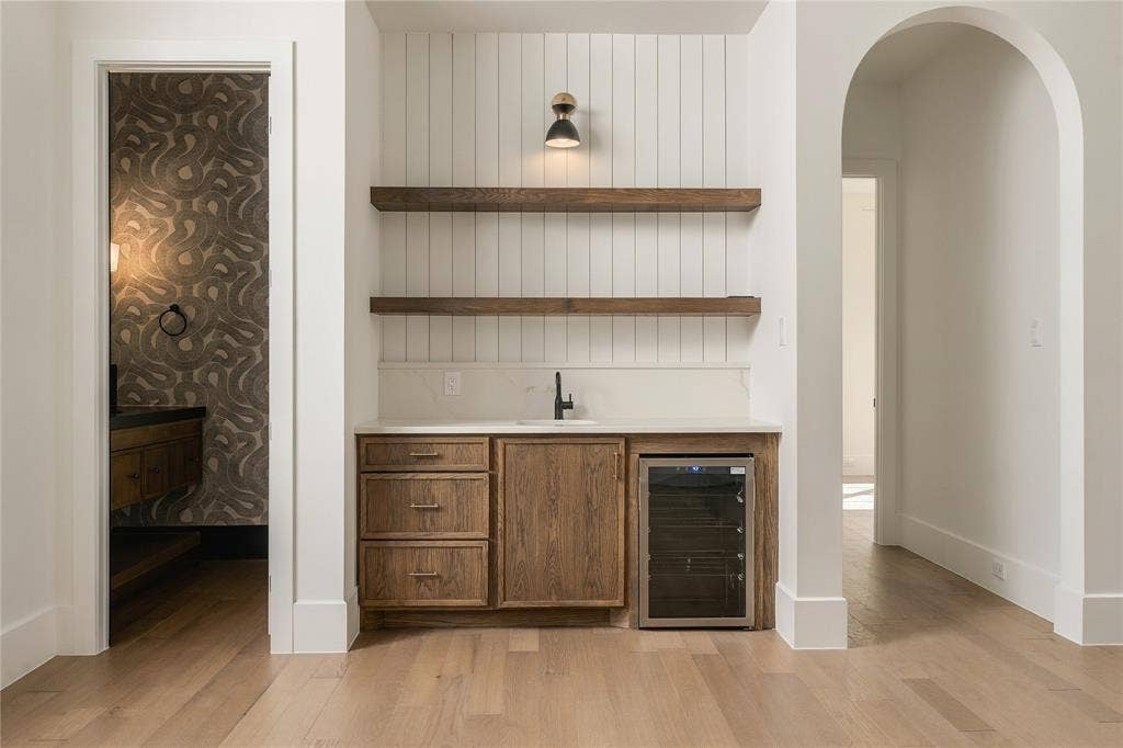Built-in wet bar featuring wood-finish cabinetry, a stone countertop, integrated wine refrigerator, vertical shiplap backsplash, and dual floating wood shelves