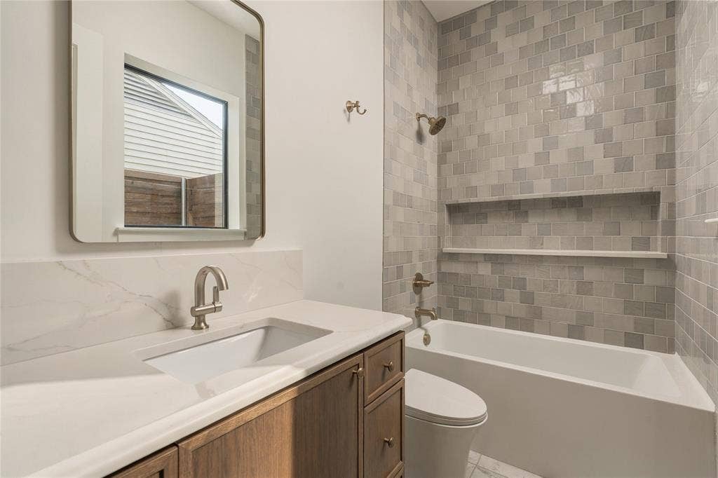 Bathroom featuring a wood-finish vanity with a stone-top integrated sink, a rectangular mirror with rounded edges, and a tub with a multi-tonal gray tile surround and integrated wall niche