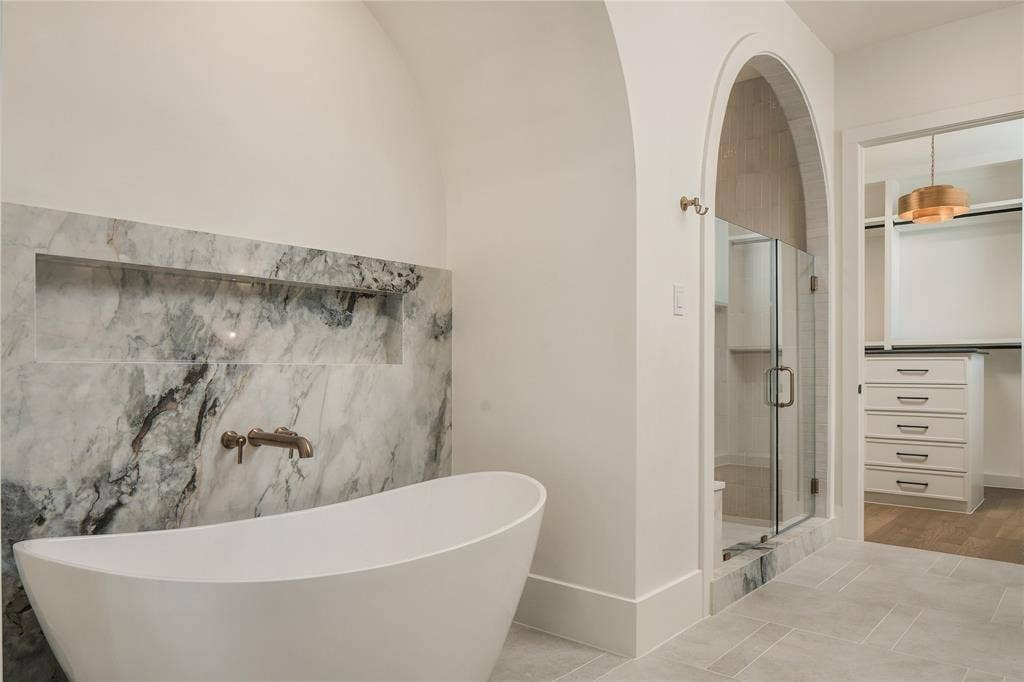 Freestanding soaking tub with wall-mounted faucet, arched shower entry, natural stone wall accent with recessed shelving, and walk-in closet featuring built-in cabinetry