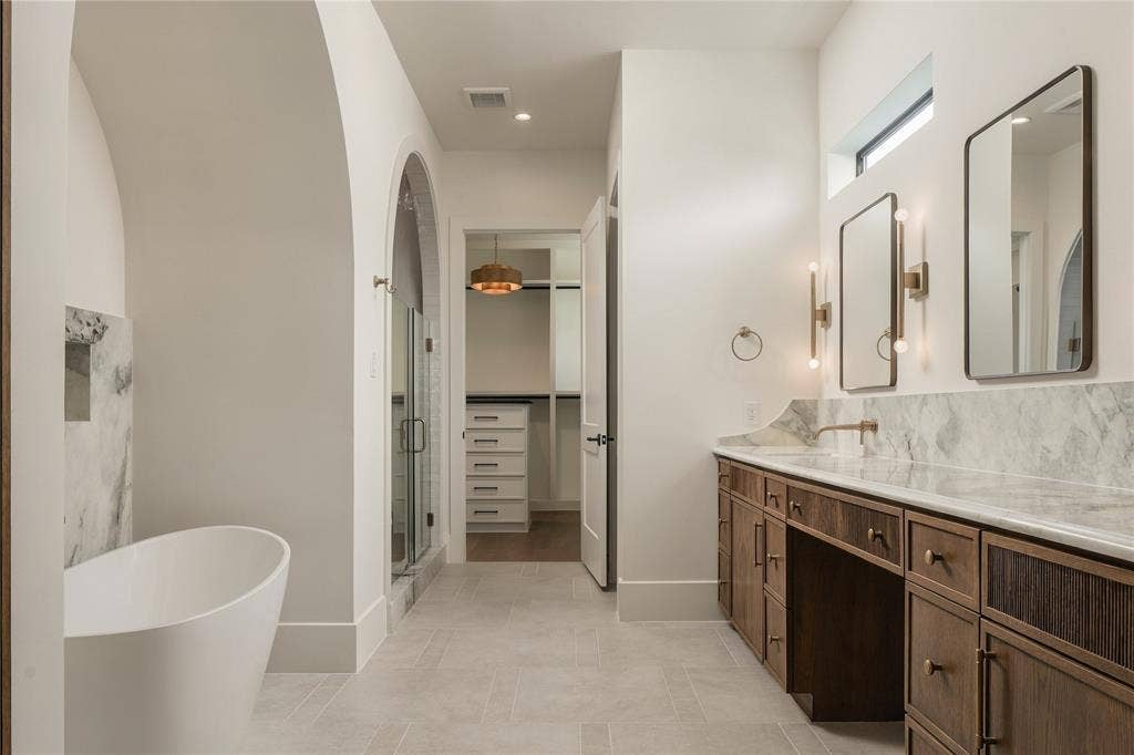 Freestanding soaking tub with a stone-accented alcove, double-vanity cabinetry with stone surfacing, arched walk-in shower, and twin rectangular mirrors with wall-mounted lighting