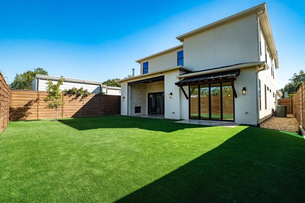 Backyard featuring synthetic turf, full-height privacy fencing, a covered patio with a built-in stone fireplace, and multi-panel glass doors
