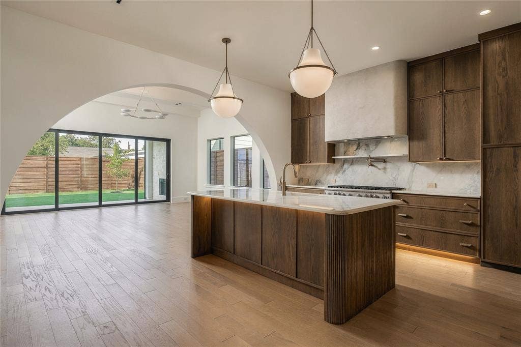 Kitchen featuring custom wood cabinetry, a stone-surround range hood, a large island with integrated under-cabinet lighting, wood-finish flooring, and a prominent architectural archway