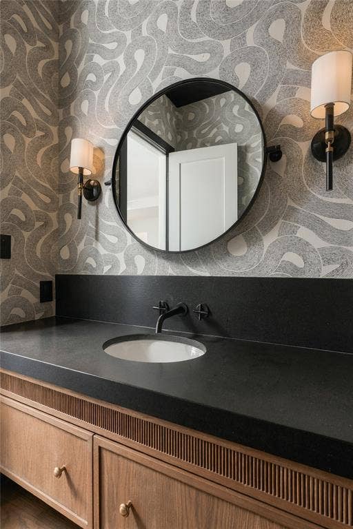 Custom wood-finish vanity with reeded detailing, dark solid-surface countertop with integrated undermount sink, circular black-frame mirror, and wall-mounted drum shade sconces
