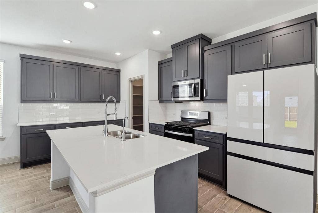 3125 Lucia Way, Princeton, TX 75407 Kitchen featuring dark shaker-style cabinetry, quartz countertops, a central island with an undermount dual-basin sink, white subway tile backsplash, and wood-finish flooring