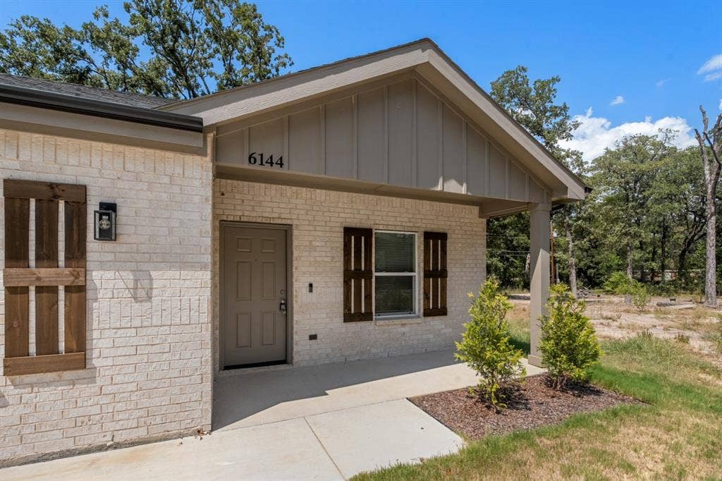 6144 Shawnee Drive, Mabank, TX 75156 Light-colored brick exterior featuring board-and-batten siding, dark wood-tone shutters, a covered concrete porch, and rectangular wall-mounted exterior lighting
