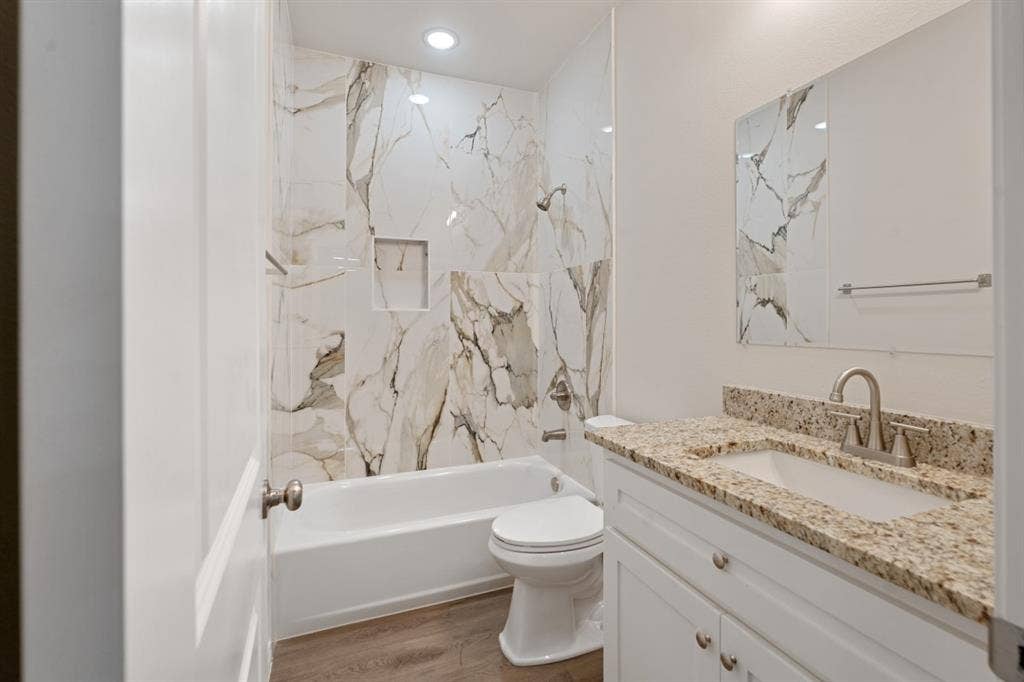 6144 Shawnee Drive, Mabank, TX 75156 Bathroom featuring a tub-shower combination with floor-to-ceiling stone-pattern tile surround, built-in wall niche, vanity with stone-surface countertop and integrated sink, and wood-finish flooring