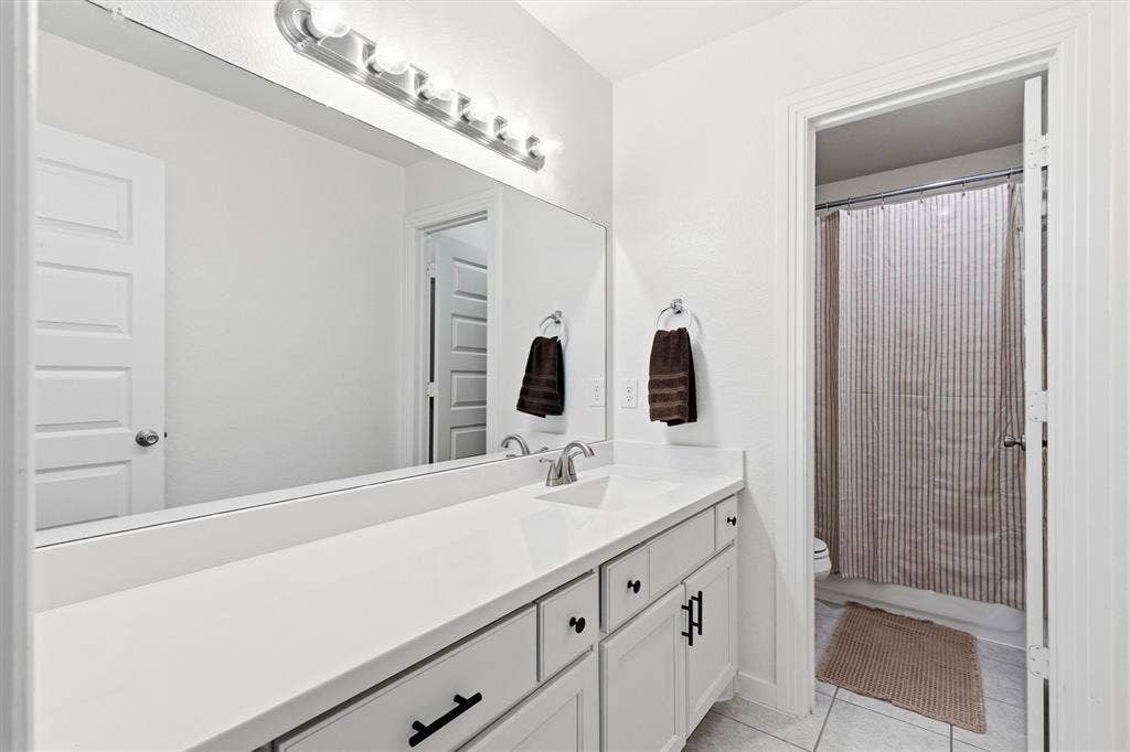 Bathroom vanity featuring white shaker-style cabinetry, matte black hardware, a solid surface countertop with an integrated sink, a full-width wall mirror, and a multi-bulb light fixture