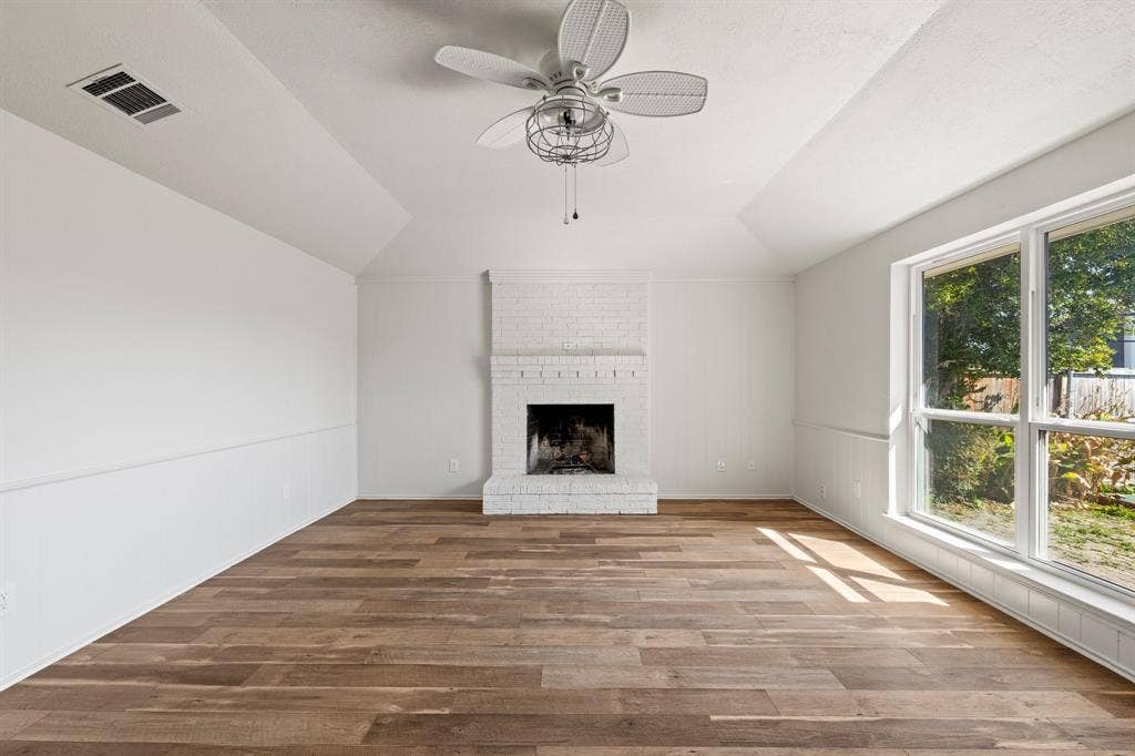 Vaulted ceiling, white-painted brick fireplace, wood-finish flooring, and large window with exterior natural light