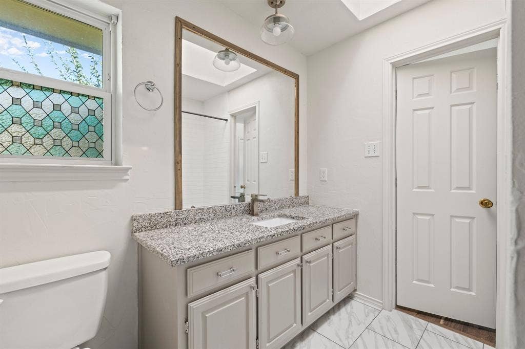 Vanity featuring a stone-look countertop, integrated sink, and shaker-style cabinetry, complemented by a large framed mirror, patterned privacy window glass, and light-toned tile flooring