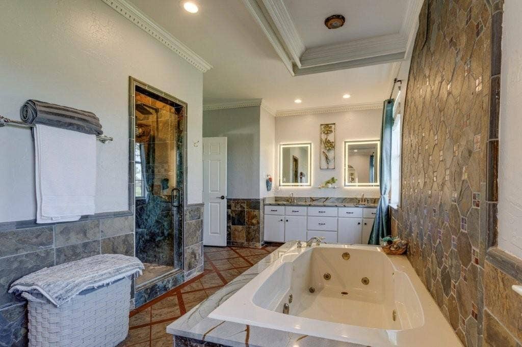Jetted soaking tub with stone surround, dual-sink vanity with illuminated mirrors, walk-in glass shower, and intricate stone wall tiling