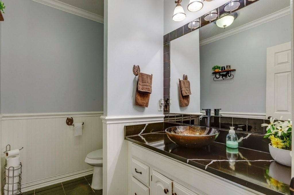 Bathroom featuring a vanity with dark stone countertop, glass vessel sink, beadboard wainscoting, and multi-bulb vanity lighting