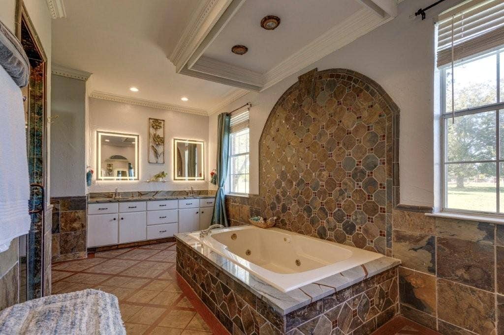 Jetted soaking tub with stone tile surround and accent wall, double vanity with illuminated mirrors, crown molding, and stone flooring