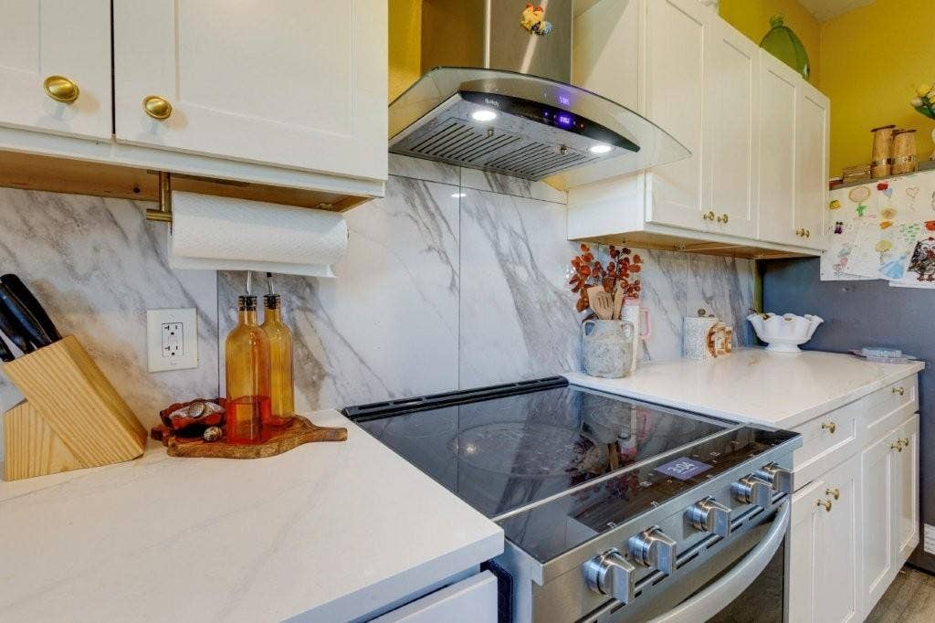 Kitchen featuring white shaker-style cabinetry, stone-finish countertops, full-height stone-look backsplash, stainless steel range, and a glass-paneled exhaust hood