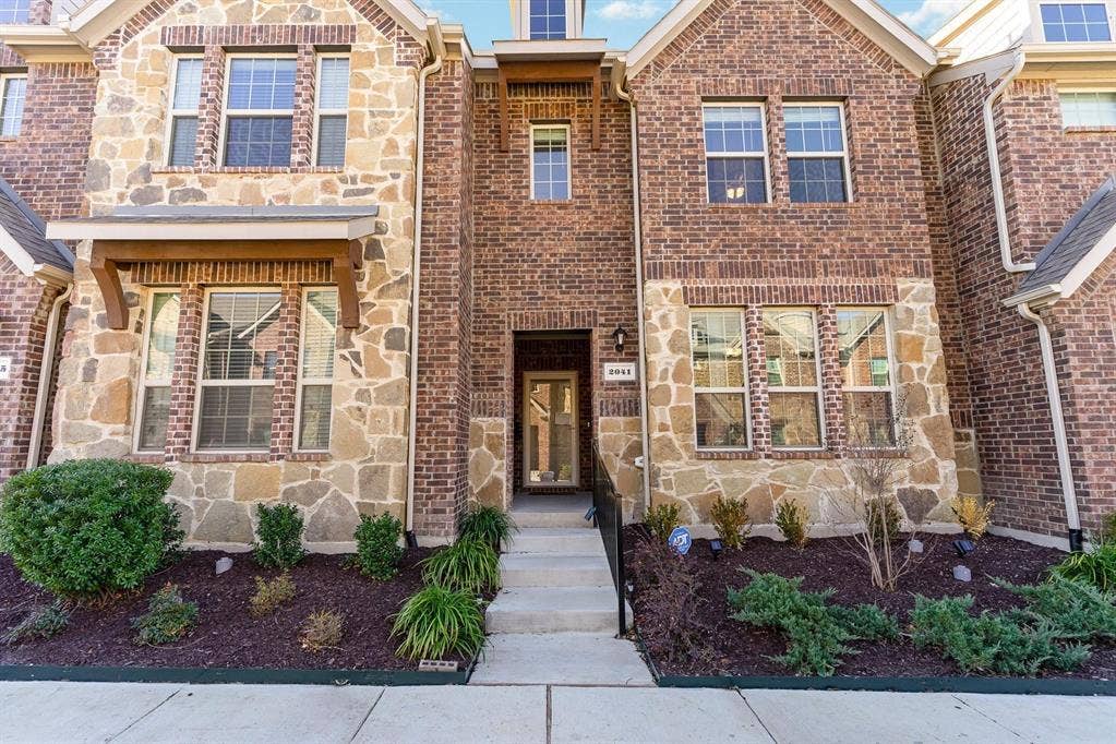 Exterior facade featuring a mix of brick and stone masonry, multi-pane windows, wood-bracketed roof accents, and concrete stairs leading to a recessed entryway